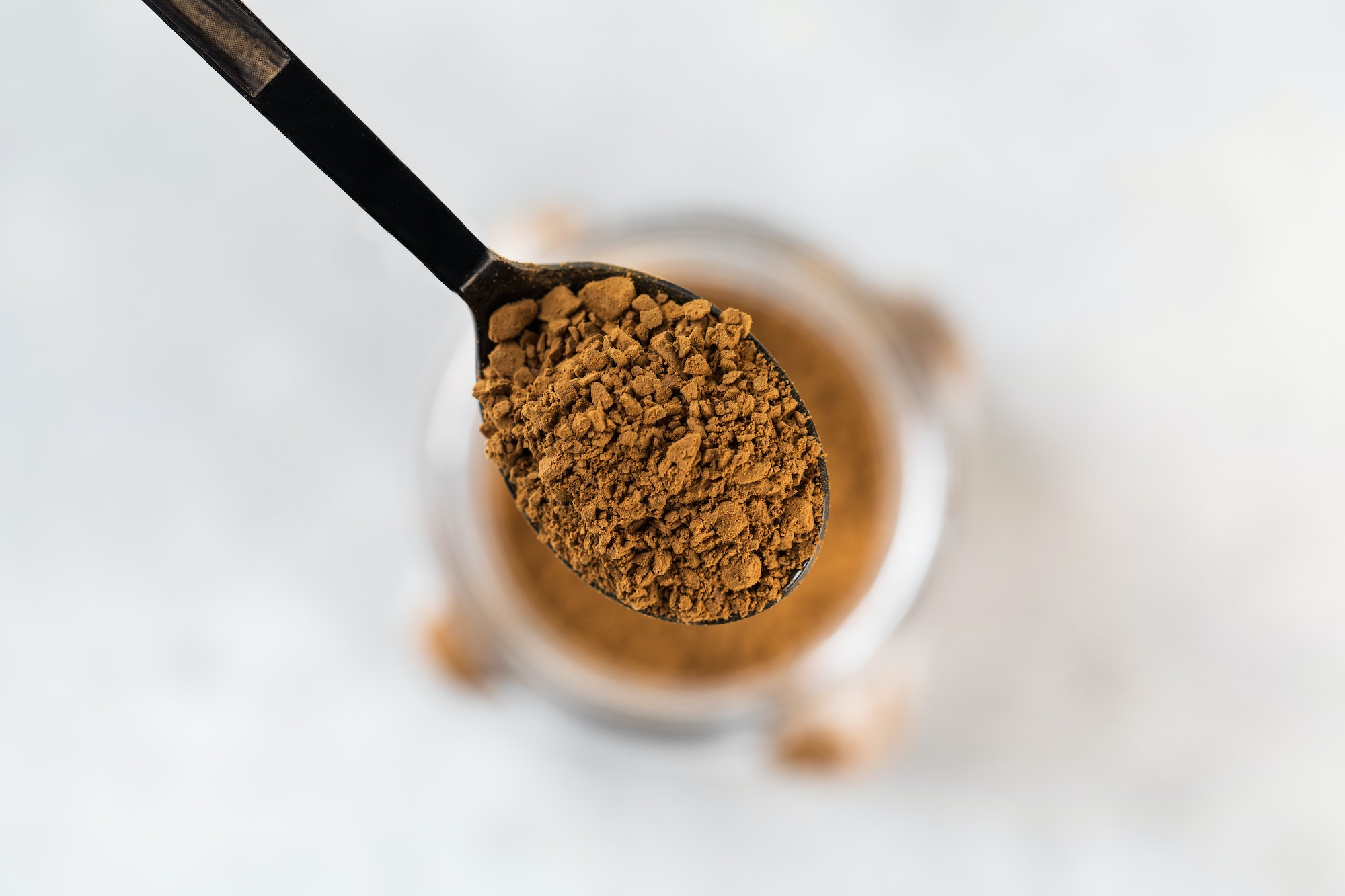 A small spoon with instant coffee granules over a glass jar of coffee.