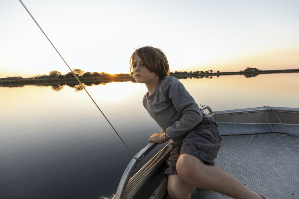 A young boy fishing from a boat on the flat calm waters of the Okavango Delta at sunset, Botswana.