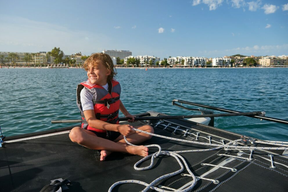 Boy with life jacket sitting in a boat holding the rudder and a rope with the shore on the